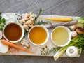 Trzy miseczki z bulionem o różnej barwie, otoczone składnikami do jego przygotowania: marchew, cebula, czosnek, imbir, grzyby, zioła. Idealne inspiracje, jak zrobić bulion.