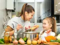 Mama i córka gotują pyszne zupy dla dzieci. Na stole warzywa: marchewka, ziemniaki, cebula, papryka i kapusta.