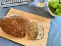 Chleb z twarogu, bogaty w ziarna i orzechy, pokrojony na desce. Obok miska zielonych winogron i szklanka wody.
