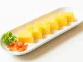 Sześć kawałków puszystego omletu tamago ułożonych na białej desce, z gałązką pietruszki i pomarańczowymi dodatkami.