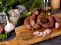 Na drewnianej desce leży pyszna, wędzona kiełbasa jałowcowa, obok czosnek, cebula i przyprawy.