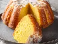 Puszysta babka na maślance, oprószona cukrem pudrem, z wyciętym kawałkiem na talerzu.
