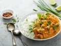 Pyszne curry masala z kurczakiem i ryżem, ozdobione szczypiorkiem i kolendrą. Obok miseczka z płatkami chili i ćwiartki limonki.