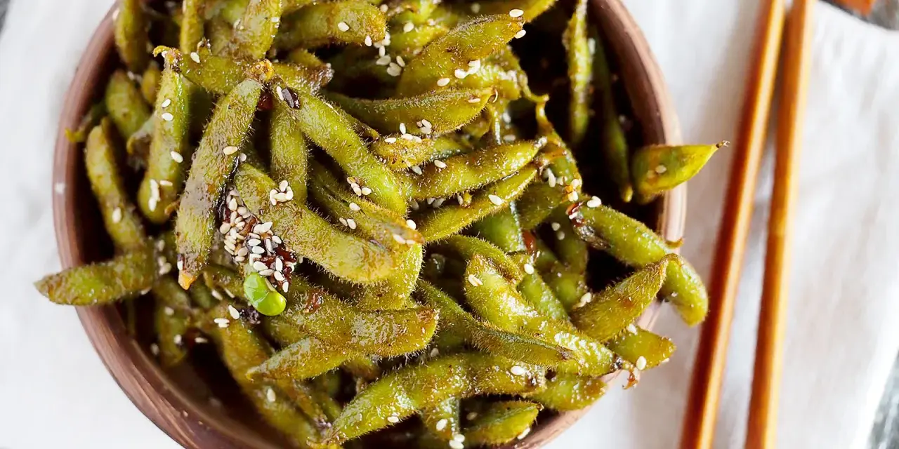 Miseczka pełna błyszczącej, przyprawionej fasolki edamame, posypanej sezamem. Obok leżą pałeczki do jedzenia.