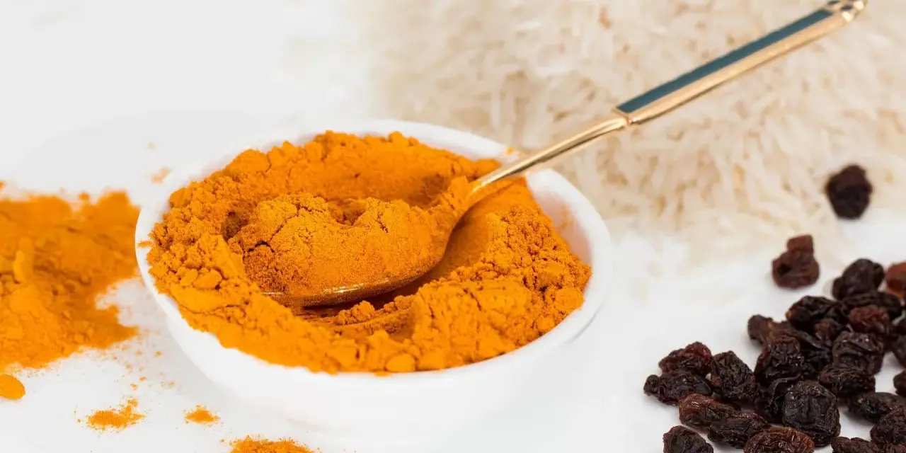 Miseczka pełna intensywnej przyprawy curry, obok ryż i rodzynki. Złota łyżeczka zanurzona w aromatycznym proszku.