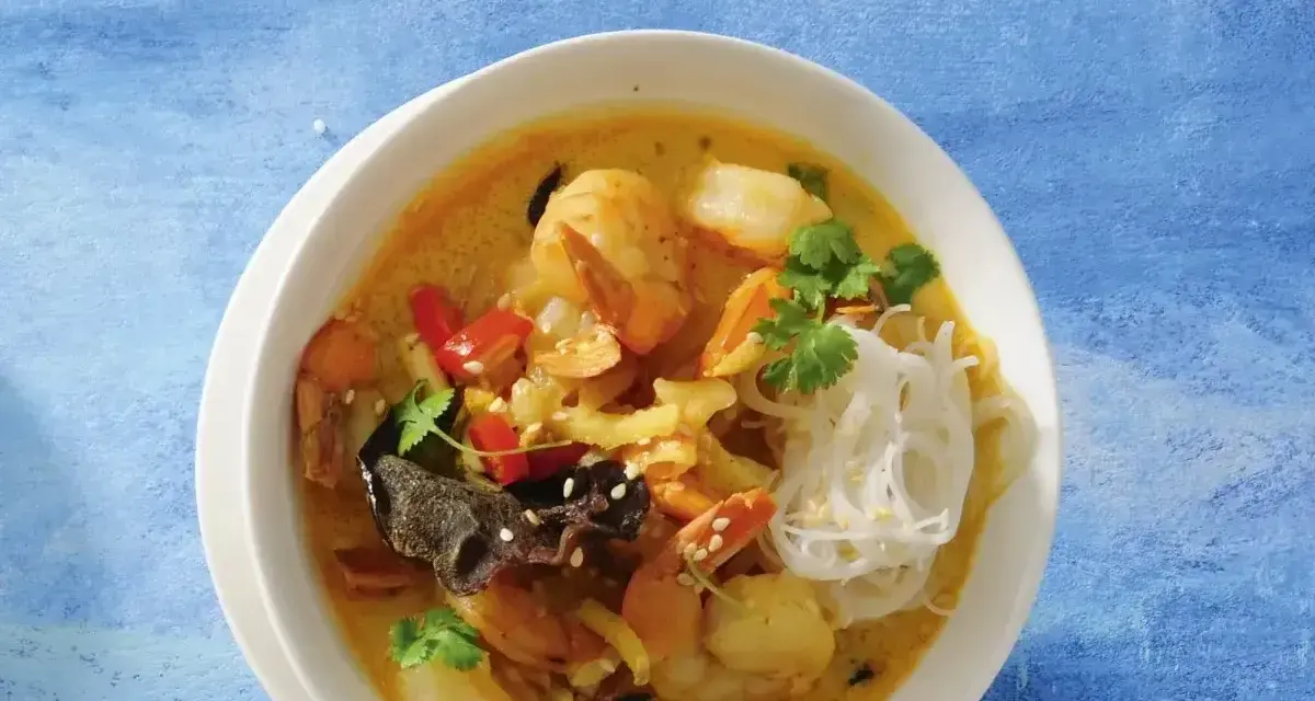 Aromatyczna zupa krewetkowa z krewetkami, makaronem ryżowym, papryczką chili i kolendrą, podana w białej misce na niebieskim tle.