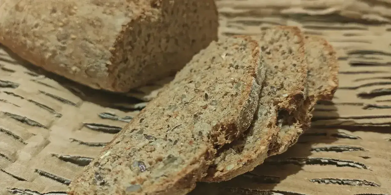 Chleb orkiszowy przepis: świeżo upieczony bochenek i kromki z ziarnami na brązowym tle.