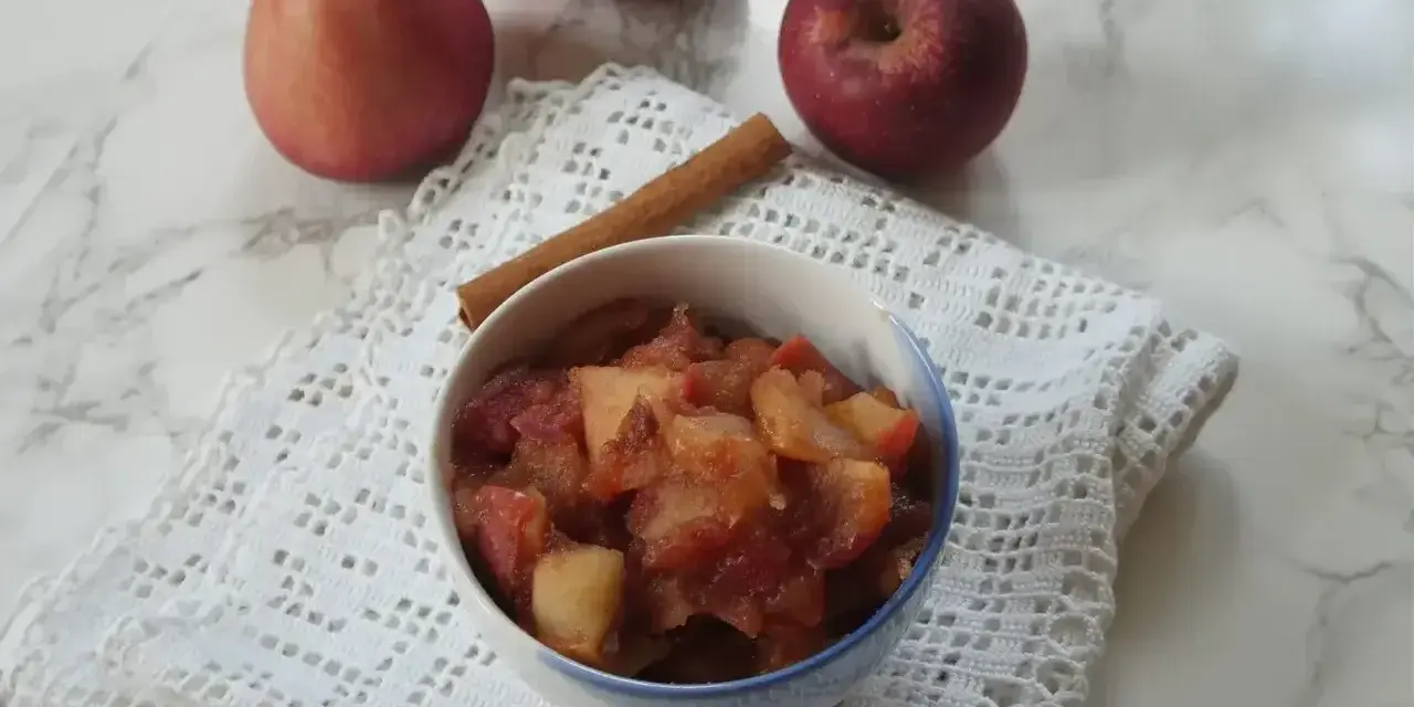Miseczka z gotowanymi jabłkami do szarlotki, obok laska cynamonu i świeże jabłka.