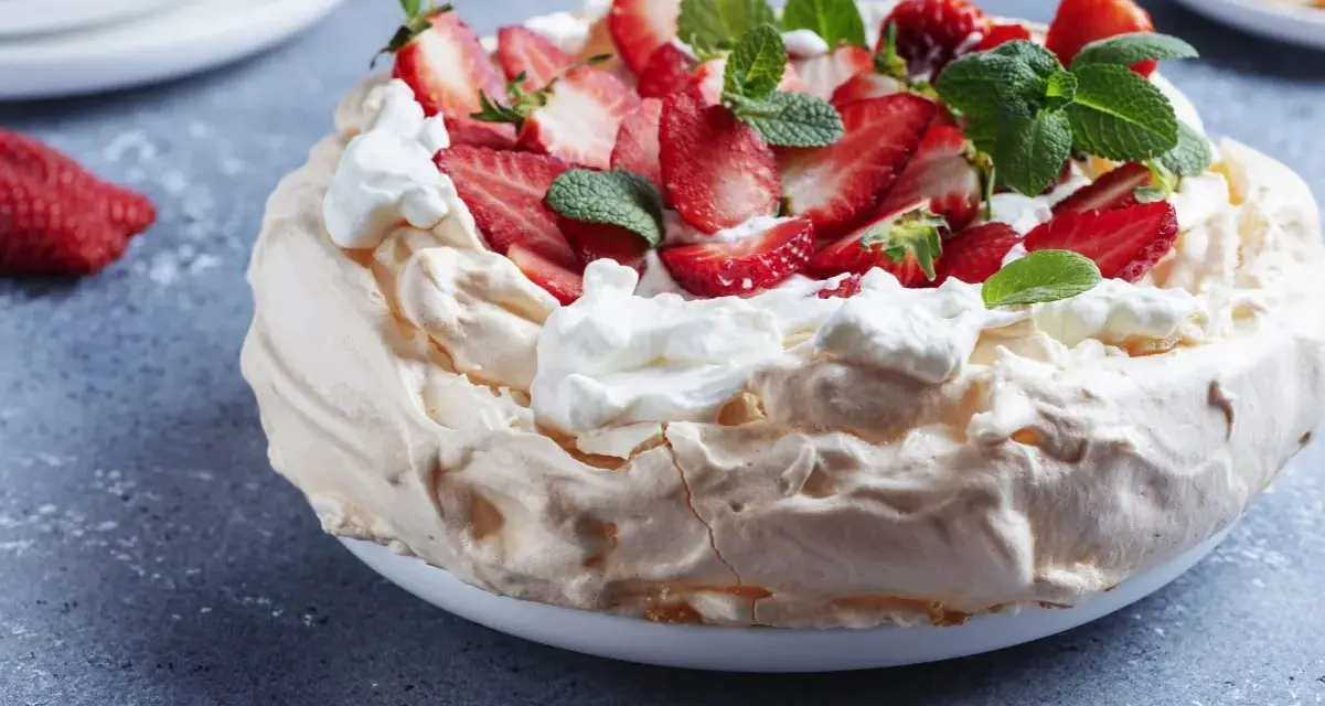 Puszysty tort bezowy z mascarpone i bitą śmietaną, ozdobiony świeżymi truskawkami i miętą.