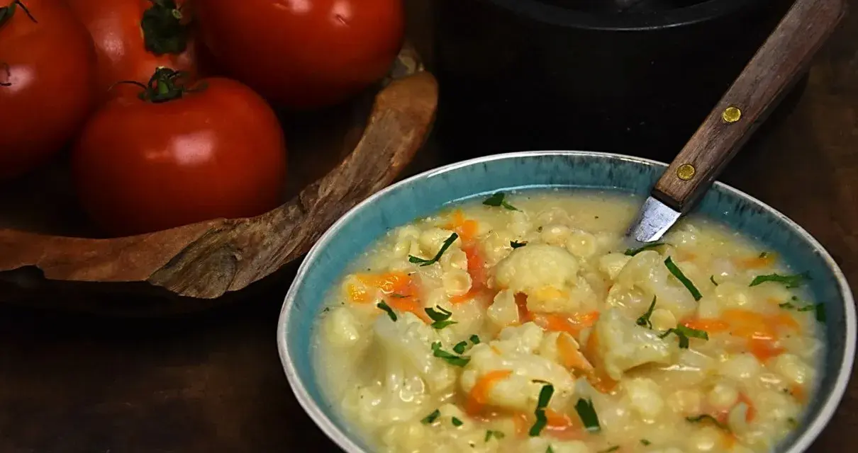 Aksamitna zupa kalafiorowa z makaronem, ozdobiona natką pietruszki. Obok miski leżą świeże pomidory.