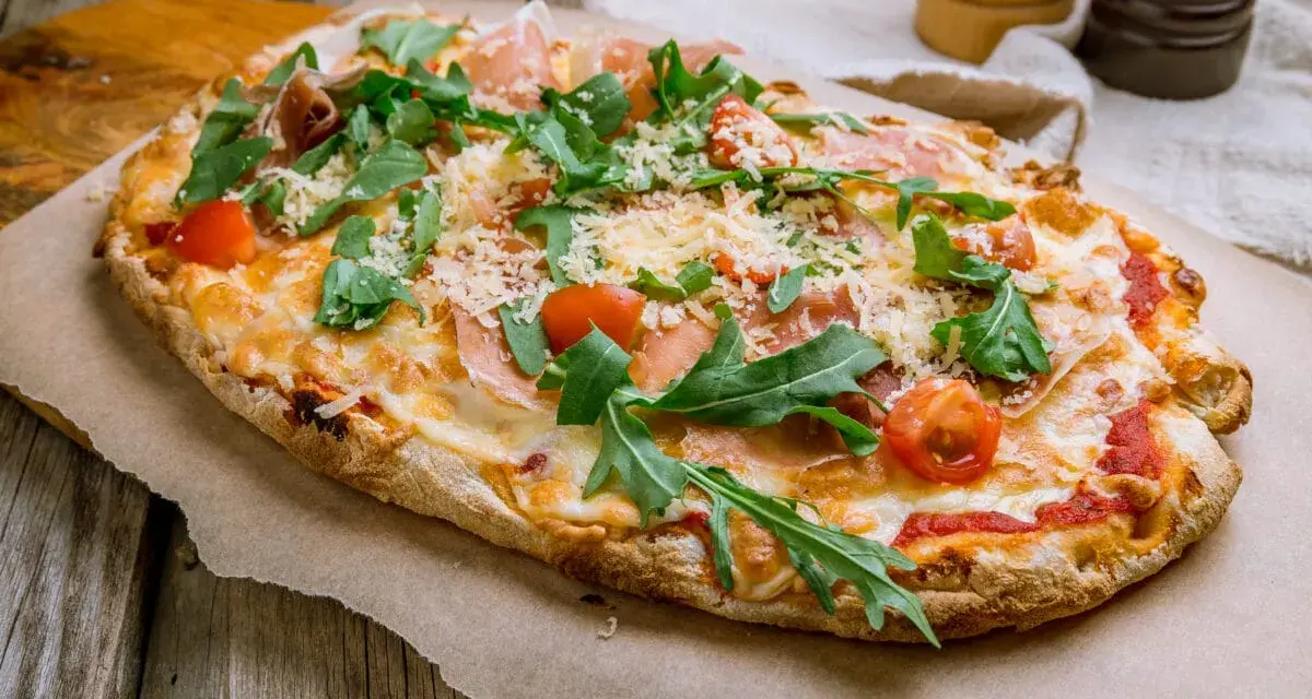 Pinsa co to? Podłużna, chrupiąca pizza z rukolą, pomidorkami i szynką parmeńską, posypana parmezanem.