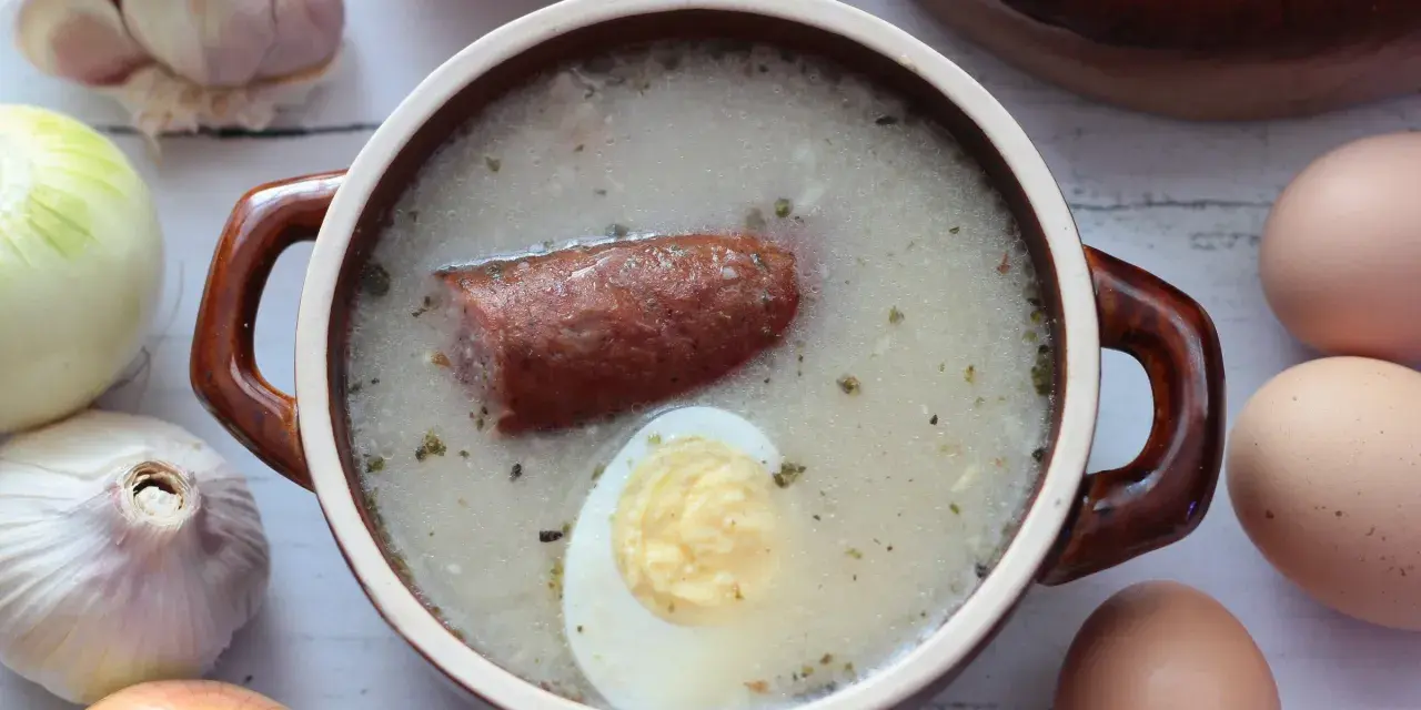 Aromaticzny żurek z kiełbasą i jajkiem, przygotowany na bazie domowego zakwasu na żurek. Obok czosnek, cebula i jajka.