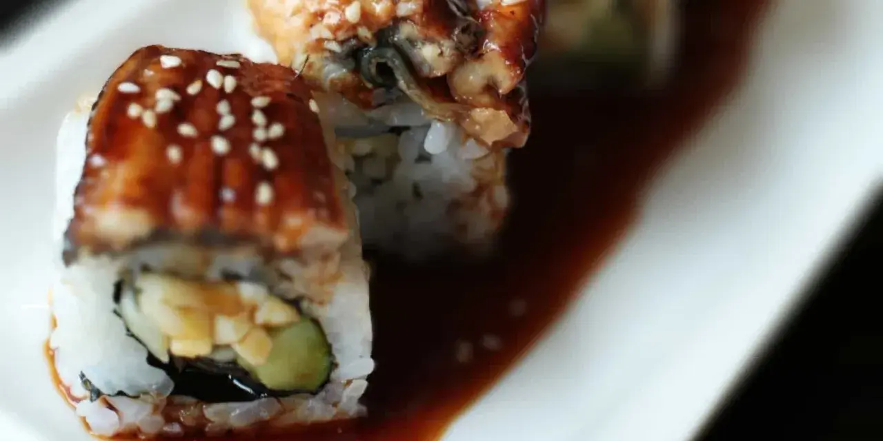 Rolka sushi z wężem morskim, polana słodkim sosem unagi i posypana sezamem, leży na talerzu.
