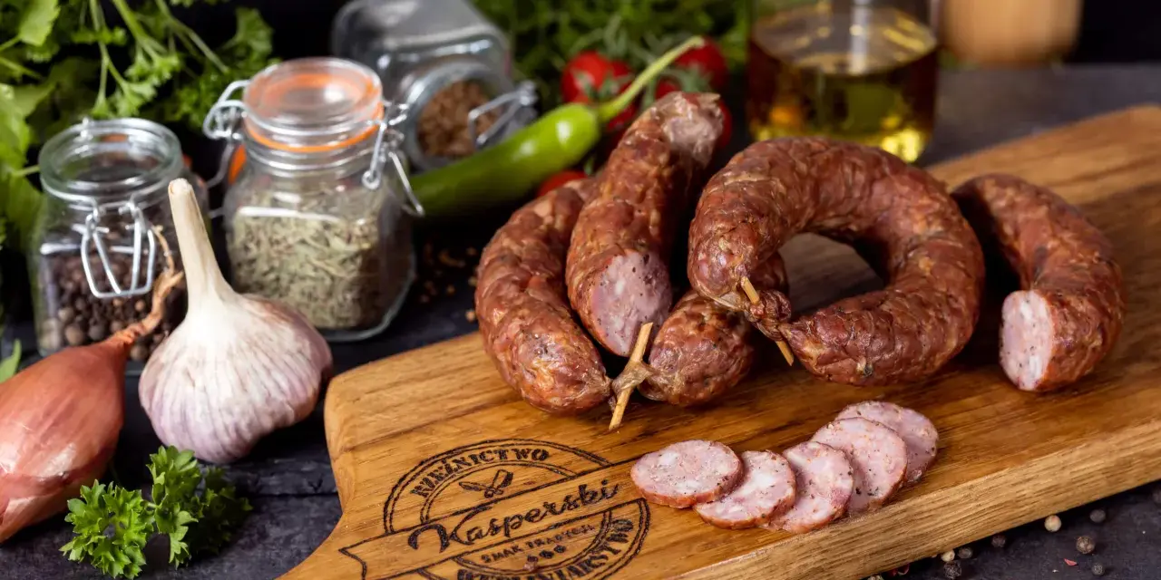 Na drewnianej desce leży pyszna, wędzona kiełbasa jałowcowa, obok czosnek, cebula i przyprawy.