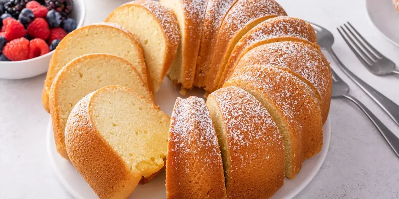 Pyszna babka piaskowa, przepis sprawdzony, posypana cukrem pudrem, obok miseczka z owocami leśnymi i widelce.