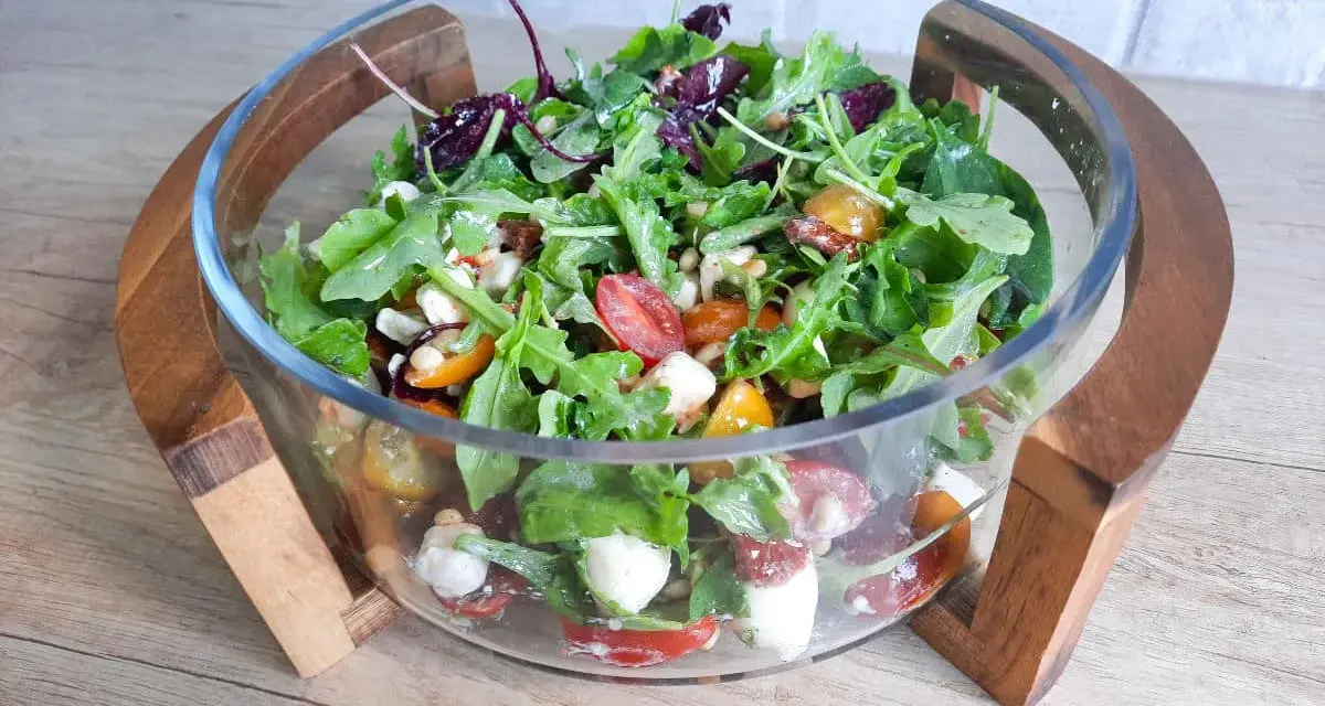 Pyszny mix sałat z pomidorkami koktajlowymi, mozzarellą i orzeszkami pinii w szklanej misie na drewnianym stojaku.