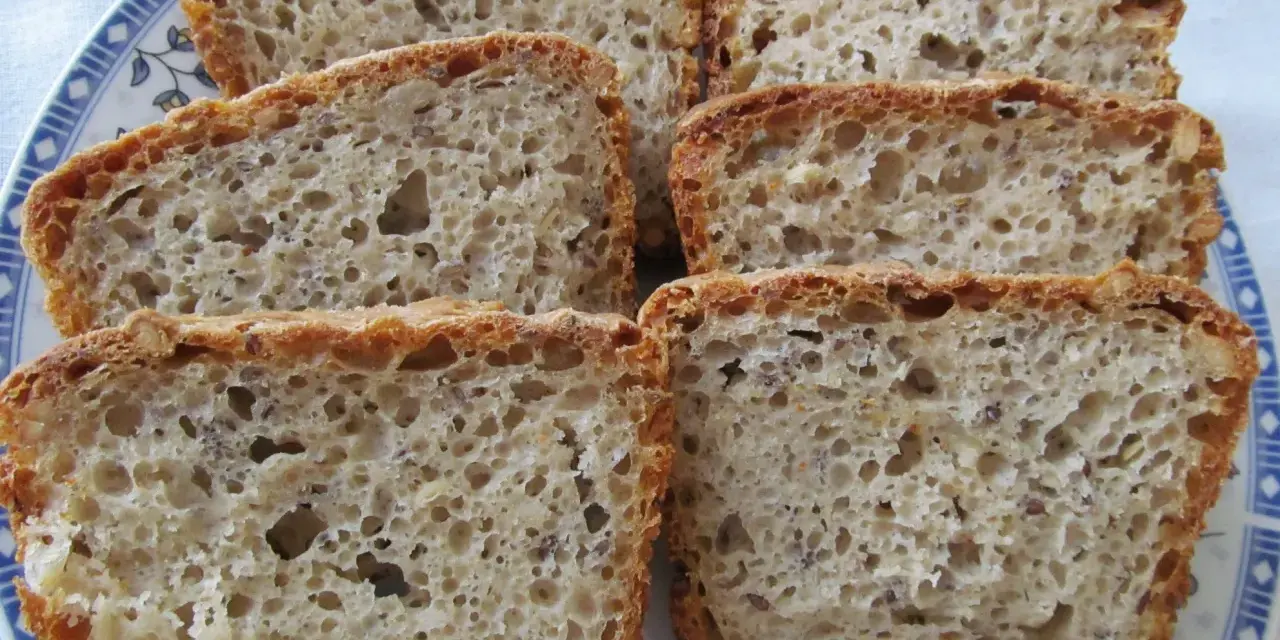 Chleb na zakwasie, pachnący jak u babci. Pokrojony w kromki, z widocznymi ziarnami, na talerzu z niebieskim wzorem.