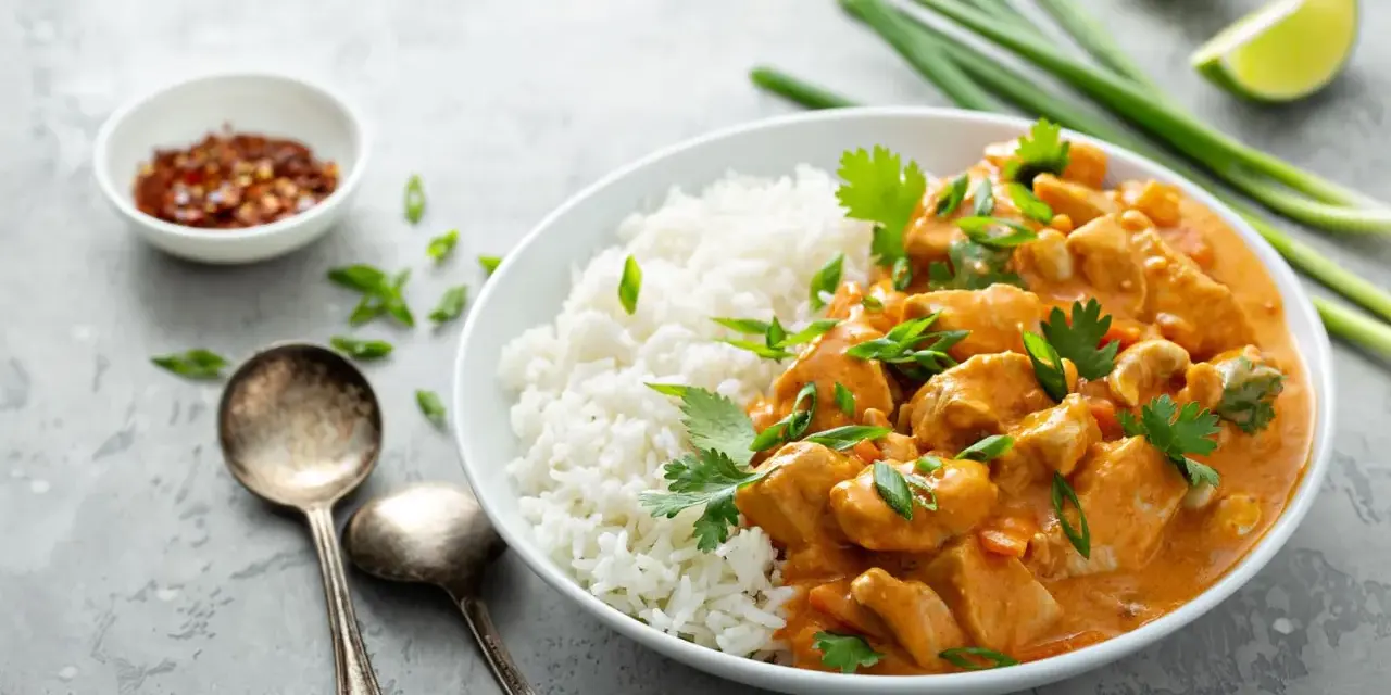 Pyszne curry masala z kurczakiem i ryżem, ozdobione szczypiorkiem i kolendrą. Obok miseczka z płatkami chili i ćwiartki limonki.