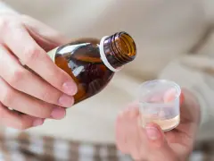 Dłoń przelewa płyn z brązowej butelki do plastikowego kieliszka. To domowy syrop na kaszel, przygotowany z naturalnych składników.