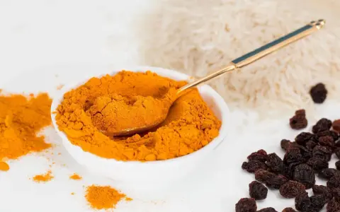 Miseczka pełna intensywnej przyprawy curry, obok ryż i rodzynki. Złota łyżeczka zanurzona w aromatycznym proszku.