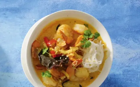 Aromatyczna zupa krewetkowa z krewetkami, makaronem ryżowym, papryczką chili i kolendrą, podana w białej misce na niebieskim tle.