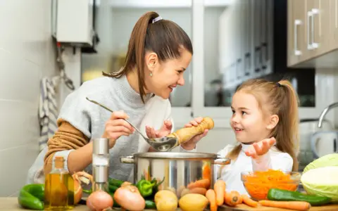 Mama i córka gotują pyszne zupy dla dzieci. Na stole warzywa: marchewka, ziemniaki, cebula, papryka i kapusta.