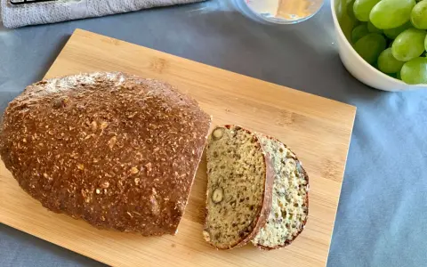 Chleb z twarogu, bogaty w ziarna i orzechy, pokrojony na desce. Obok miska zielonych winogron i szklanka wody.