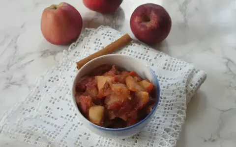 Miseczka z gotowanymi jabłkami do szarlotki, obok laska cynamonu i świeże jabłka.