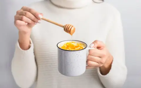 Ręce w białym swetrze dodają miód do kubka z mlekiem. Idealny napój na chłodne dni.