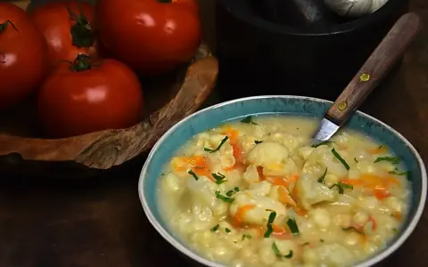 Aksamitna zupa kalafiorowa z makaronem, ozdobiona natką pietruszki. Obok miski leżą świeże pomidory.