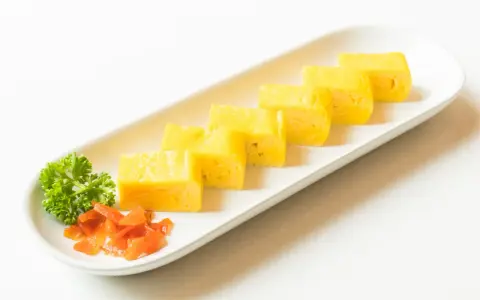 Sześć kawałków puszystego omletu tamago ułożonych na białej desce, z gałązką pietruszki i pomarańczowymi dodatkami.
