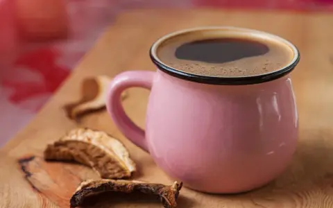 Różowy kubek z aromatyczną kawą grzybową na drewnianej desce, obok suszone grzyby.