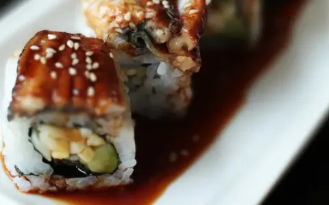 Rolka sushi z wężem morskim, polana słodkim sosem unagi i posypana sezamem, leży na talerzu.