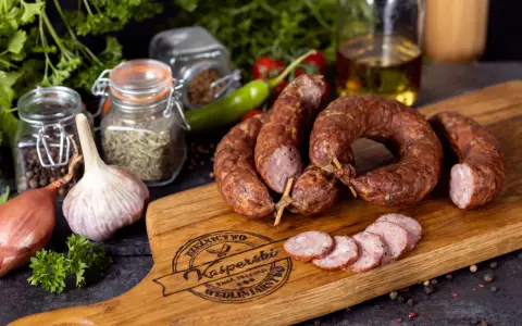 Na drewnianej desce leży pyszna, wędzona kiełbasa jałowcowa, obok czosnek, cebula i przyprawy.