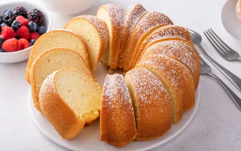 Pyszna babka piaskowa, przepis sprawdzony, posypana cukrem pudrem, obok miseczka z owocami leśnymi i widelce.