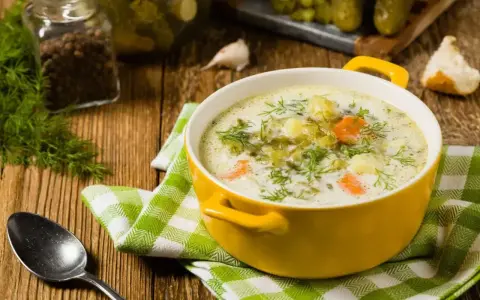 Zupa ogórkowa z koperkiem, ziemniakami i marchewką. W tle słoik z pieprzem, czosnek i ogórki kiszone. Idealne jakie przyprawy do zupy ogórkowej?