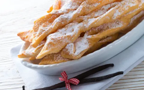 Chrupiące faworki, przepis babci, posypane cukrem pudrem, obok laski wanilii i czerwono-biała kokardka.
