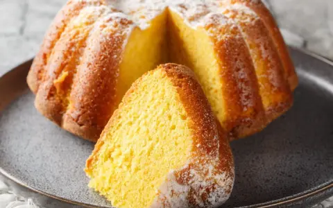 Puszysta babka na maślance, oprószona cukrem pudrem, z wyciętym kawałkiem na talerzu.