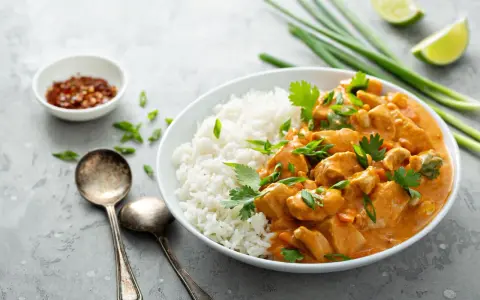 Pyszne curry masala z kurczakiem i ryżem, ozdobione szczypiorkiem i kolendrą. Obok miseczka z płatkami chili i ćwiartki limonki.