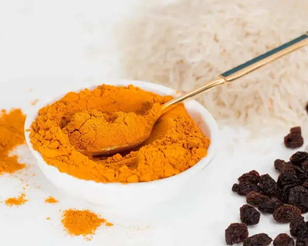 Miseczka pełna intensywnej przyprawy curry, obok ryż i rodzynki. Złota łyżeczka zanurzona w aromatycznym proszku.