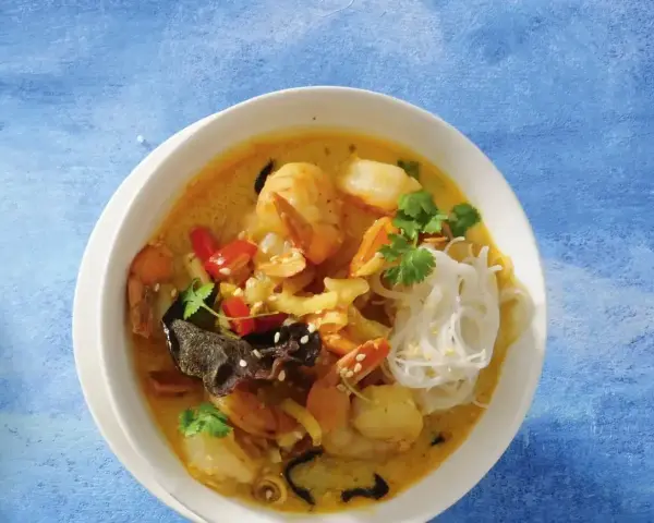 Aromatyczna zupa krewetkowa z krewetkami, makaronem ryżowym, papryczką chili i kolendrą, podana w białej misce na niebieskim tle.