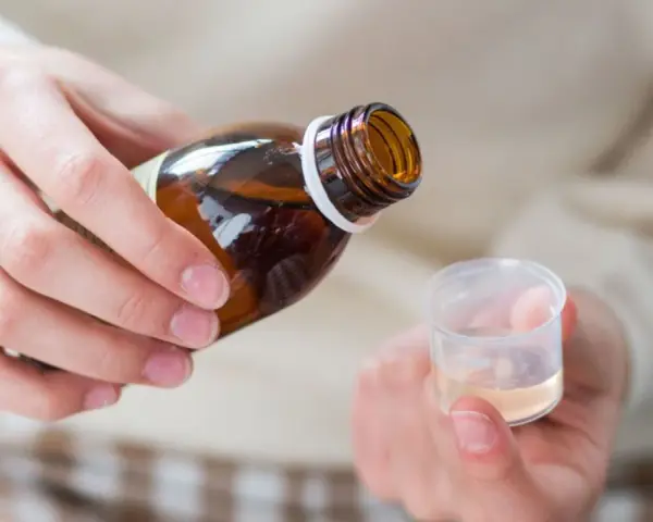 Dłoń przelewa płyn z brązowej butelki do plastikowego kieliszka. To domowy syrop na kaszel, przygotowany z naturalnych składników.