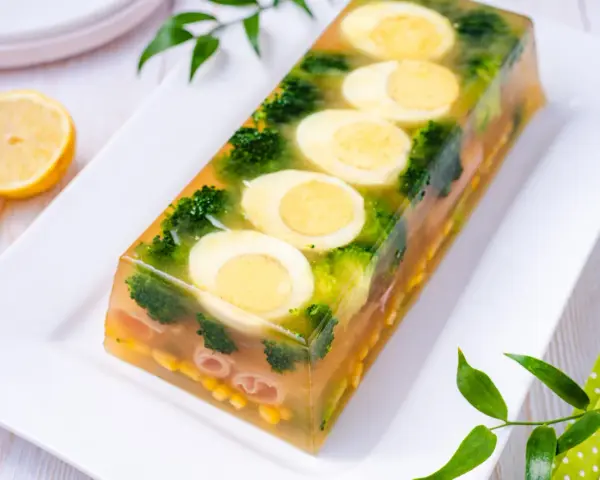 Eleganckie jajka w galarecie z brokułami i kukurydzą, ozdobione plasterkami cytryny.