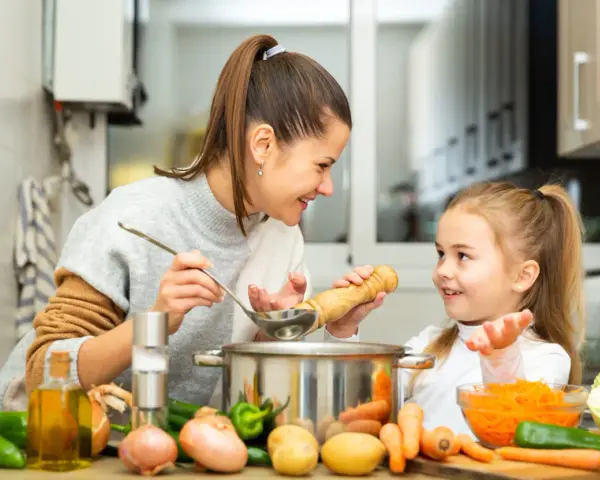 Mama i córka gotują pyszne zupy dla dzieci. Na stole warzywa: marchewka, ziemniaki, cebula, papryka i kapusta.