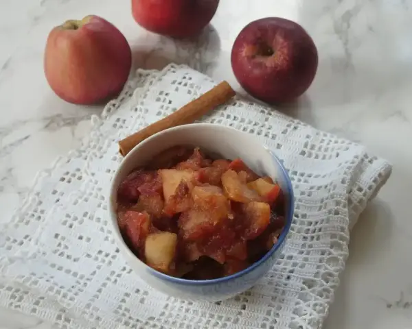 Miseczka z gotowanymi jabłkami do szarlotki, obok laska cynamonu i świeże jabłka.