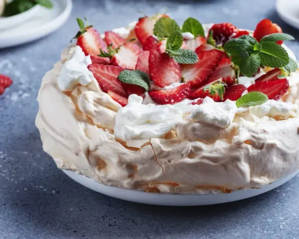 Puszysty tort bezowy z mascarpone i bitą śmietaną, ozdobiony świeżymi truskawkami i miętą.