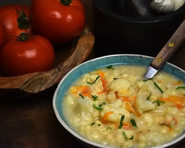 Aksamitna zupa kalafiorowa z makaronem, ozdobiona natką pietruszki. Obok miski leżą świeże pomidory.