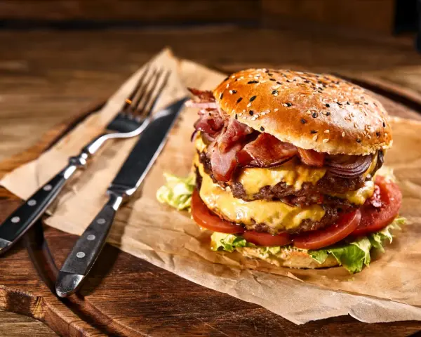 Podwójny burger z soczystym mięsem na burgery, serem, bekonem i warzywami, gotowy do zjedzenia z widelcem i nożem.