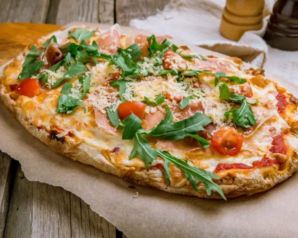 Pinsa co to? Podłużna, chrupiąca pizza z rukolą, pomidorkami i szynką parmeńską, posypana parmezanem.