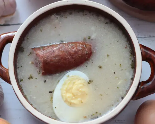 Aromaticzny żurek z kiełbasą i jajkiem, przygotowany na bazie domowego zakwasu na żurek. Obok czosnek, cebula i jajka.